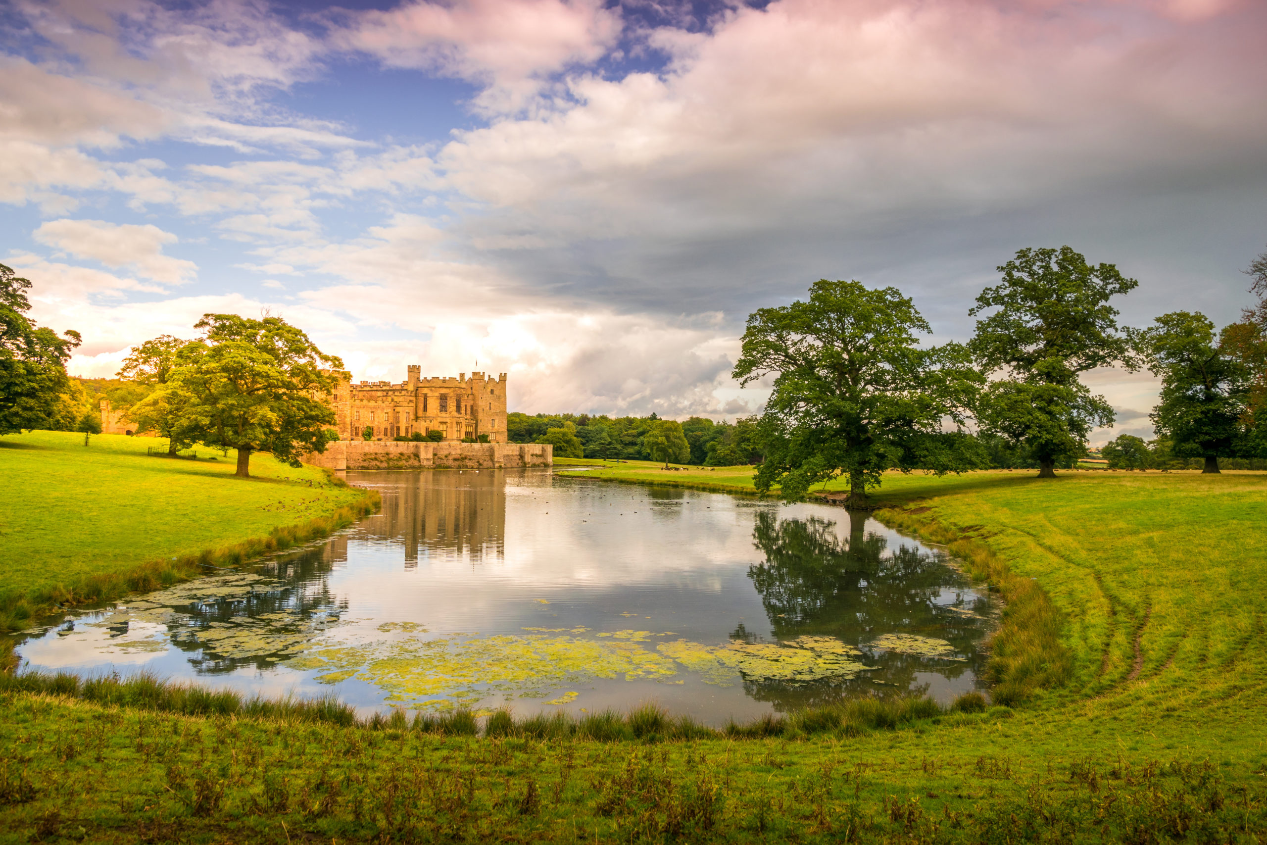 Raby Castle reflected in a pond and a parklike setting.